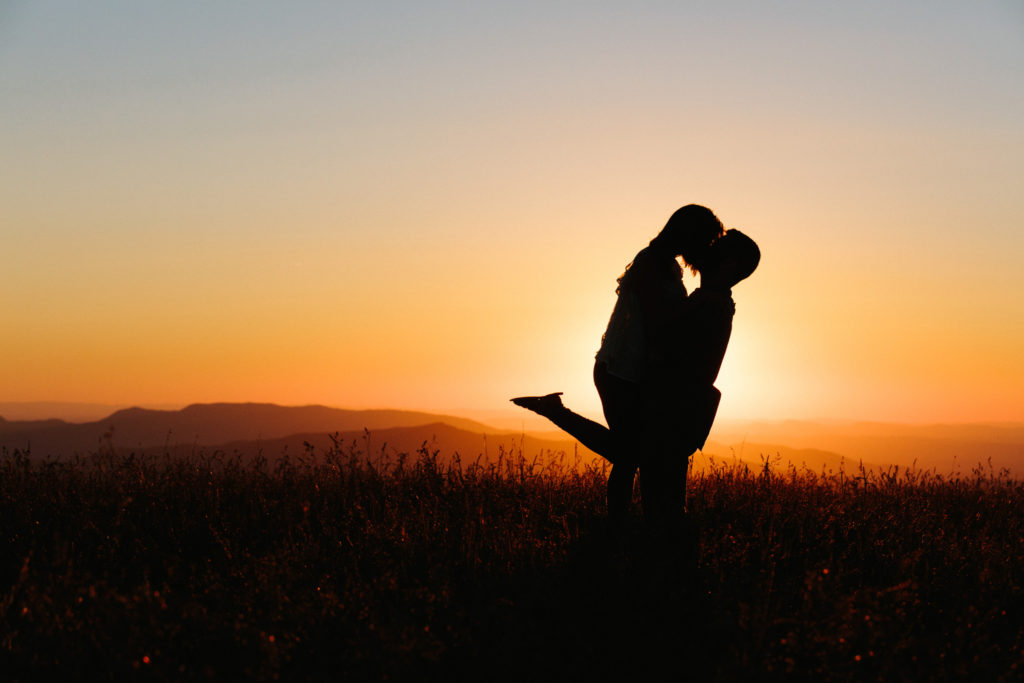Photo of Jenna and Jim on top of Max Patch, they are back lite standing in tall grass with the sun setting directly behind them on this Adventure Engagement Photography session