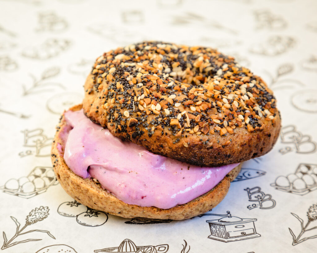 Fig & Sorghum bagel sitting on white parch paper that has designs of eggs, wheat plants, fish, and kitchen tools. This bagel is called Sweet Appalachia and has what appear to be rich pink cream cheese on a large poppy seeded bagel.  It listed ingredients are homemade jam, fig, sorghum, and cream cheese. 