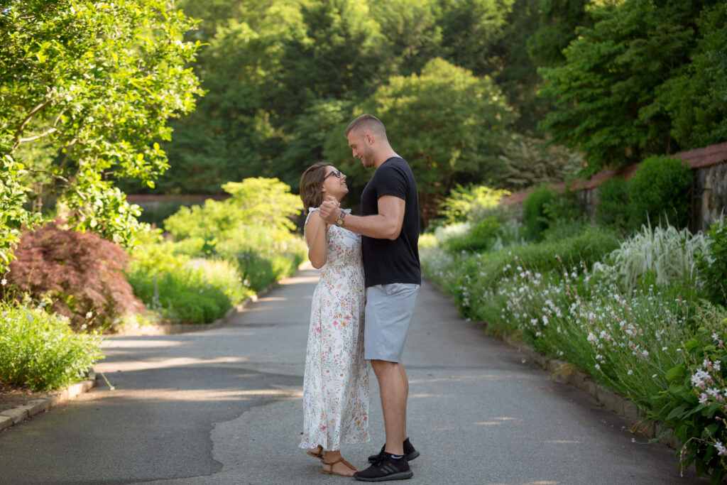 The newly engaged couple celebrating their surprise wedding proposal by dancing in the garden. Rows of flowers line the area in which they are dancing.