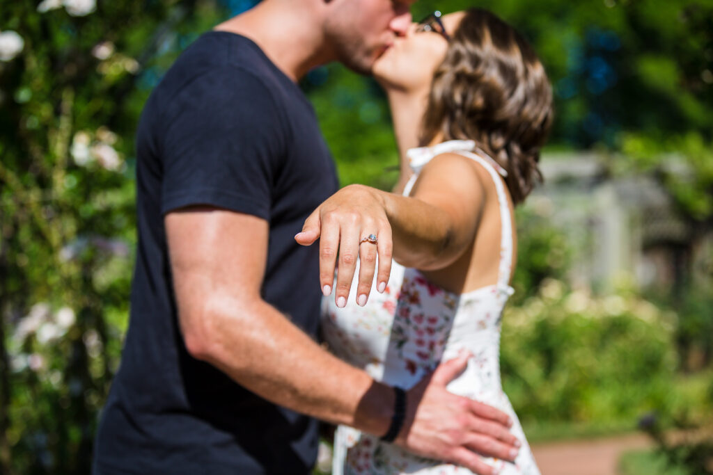 Picture of engagement ring on her hand, the ring is in focus while the couple are blurred out in the background kissing. 