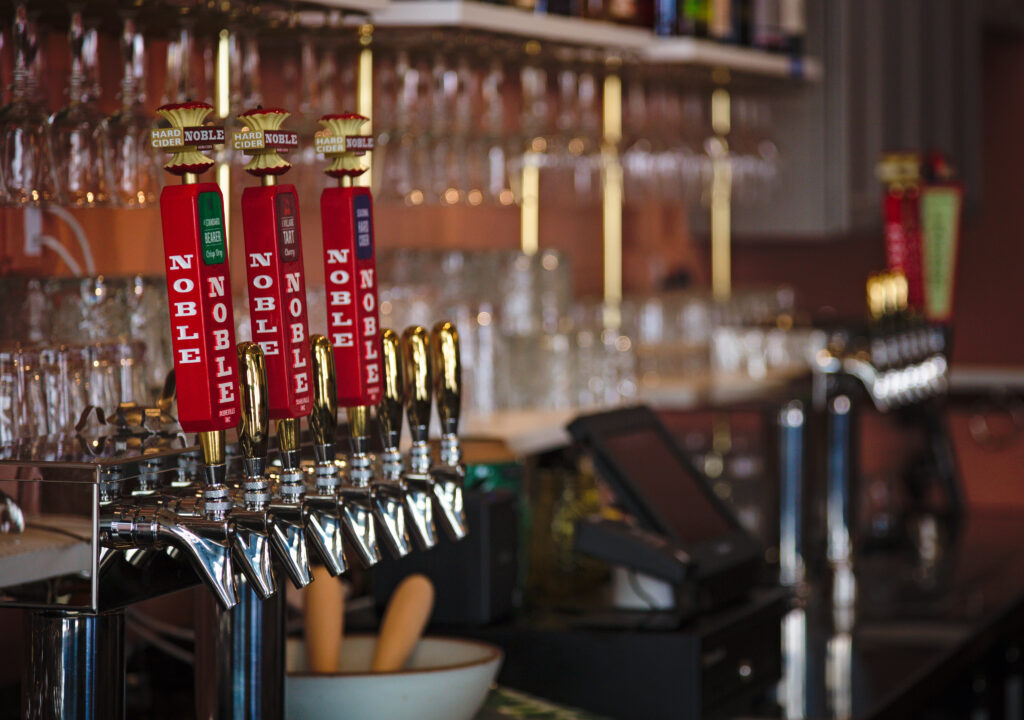 Photo looking at the taps at Noble Cidery. You can see bar glasses blurred in the background