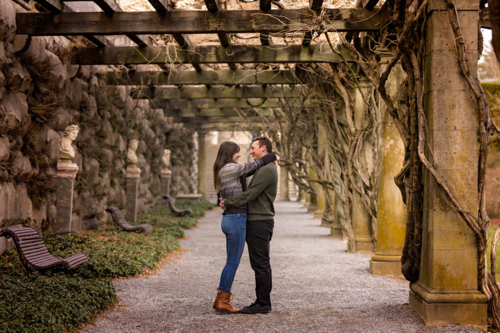 Photo of the couple holding one another under the Pergola at Biltmore Estate in Asheville it is winter, so the Wisteria is dead; however the vines remain in creating a dramatic visual effect.