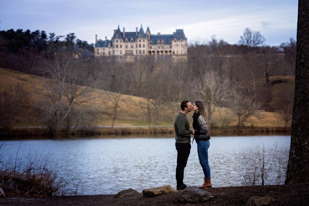 The couple standing on the edge of the Lagoon at Biltmore Estate in Asheville North Carolina, with the Biltmore looming on the hill in the distant background. It is clear from this engagement photo that it was shot during the winter has all the leaves have fallen from the tree.
