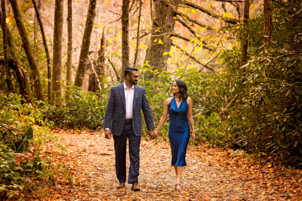The couple is walking, holding hands, staring intensely into one another's eyes. The hiking trail is covered golden red beautiful Hughes from the Fall colors that have dropped into the forest floor.