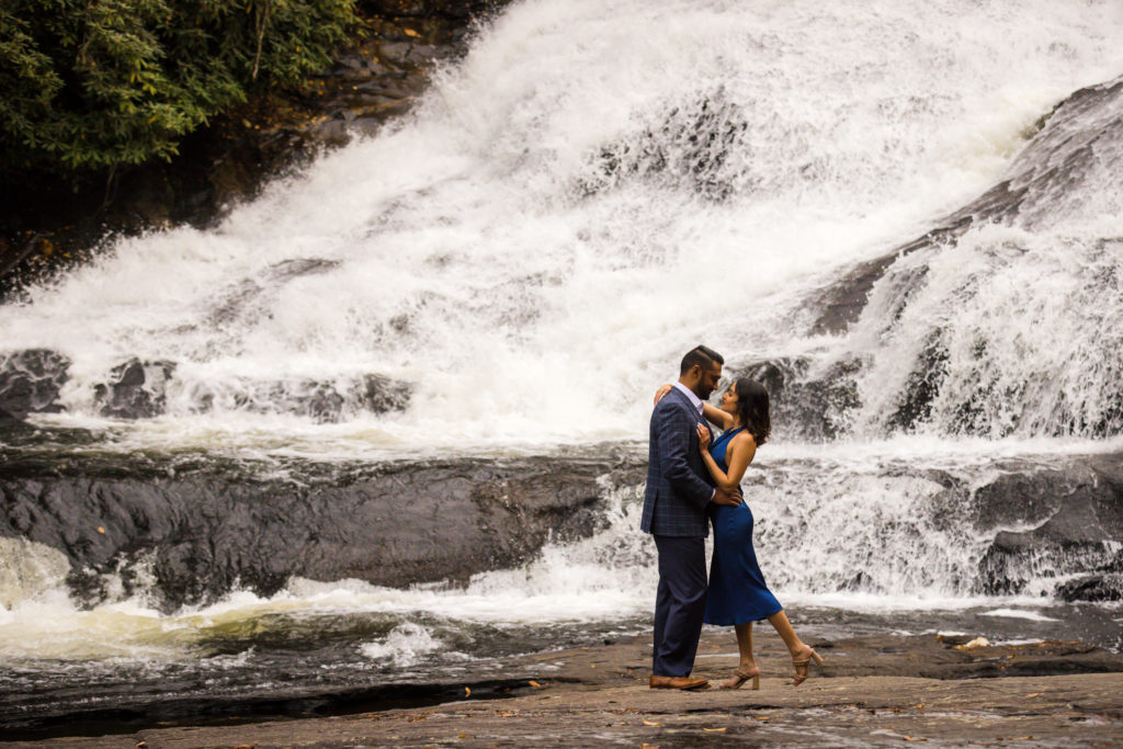 Alok and Sunaina are standing at the base of Triple Falls at DuPont State Recreational Forest, holding one another with the water rushing in the background.