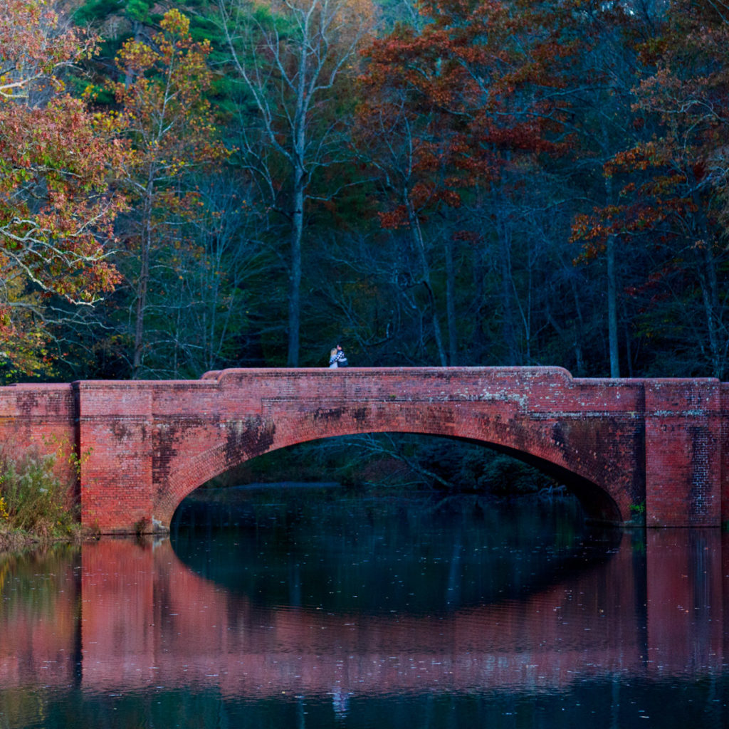 Photo from across the Biltmore bass pond oh, this is the bridge that leads across the best part. It is made of brick and glistens red and in Setting Sun there is a reflection in the  pond of the bridge and the couple