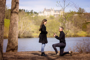 Surprise proposal photography at the Biltmore Estate, Matt is down on one knee with the Lagoon in the background and the Biltmore Estate on the hill. The ring is in his hand and he is proposing to Meredith.