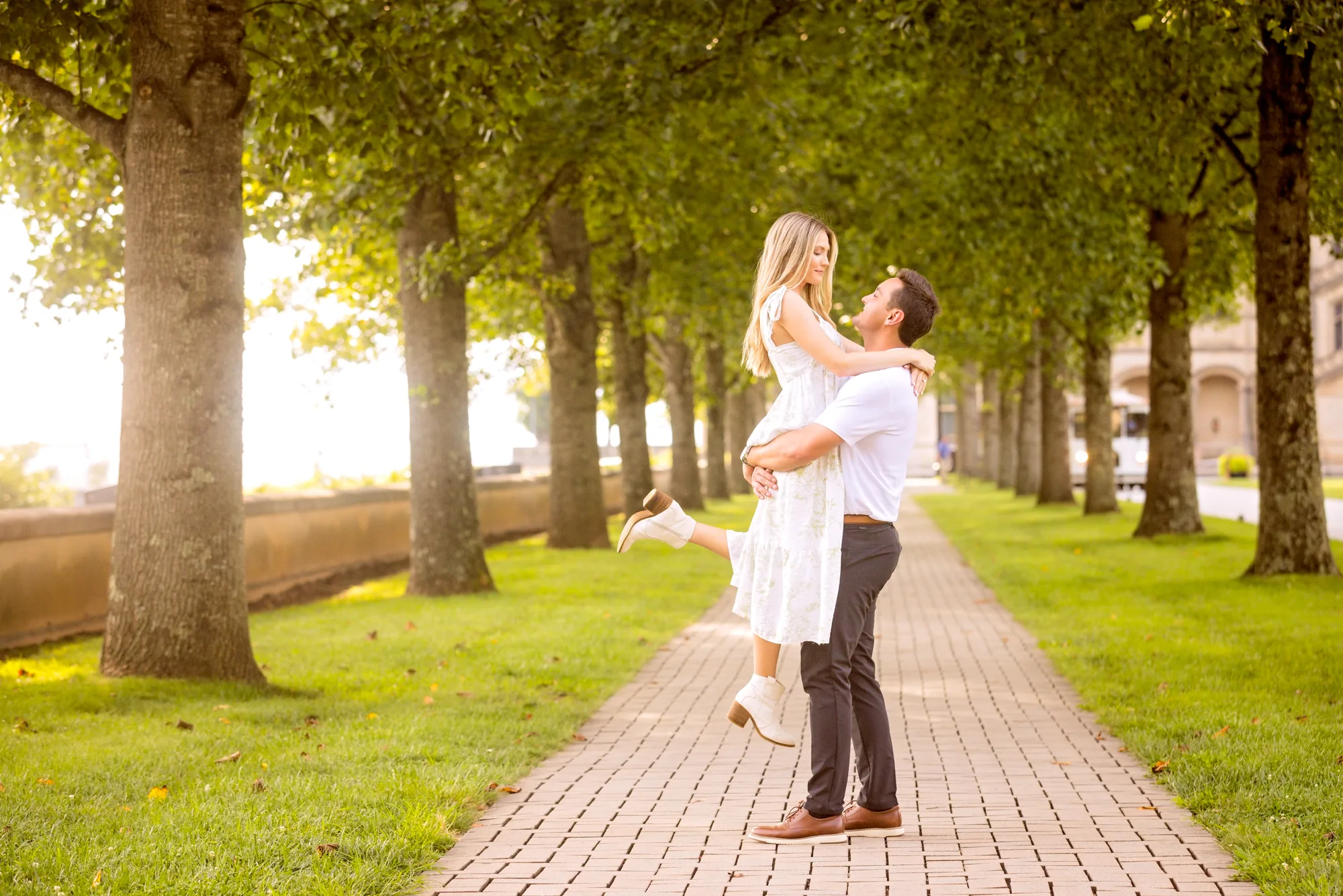 Alex lifts Andrea off the brick path in the tree-lined allée at Biltmore Estate, Andrea laughing in a white floral sundress and white ankle boots with her arms around his neck, golden sun flaring through the rows of trees behind them, a storybook engagement photo in Asheville NC