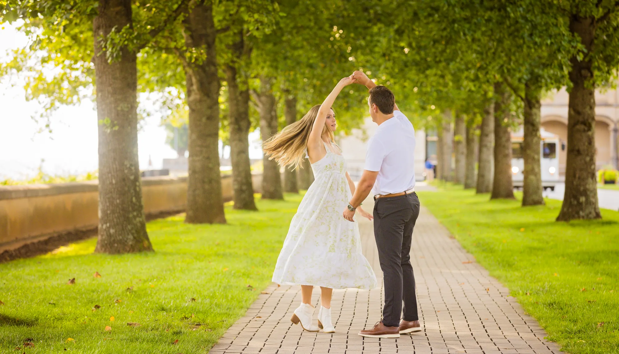 Alex twirls Andrea under his arm on the brick walkway of the allée at Biltmore Estate, her long blonde hair and white floral dress spinning in golden backlight beneath the double row of trees, a dreamy sun-drenched engagement photo in Asheville NC