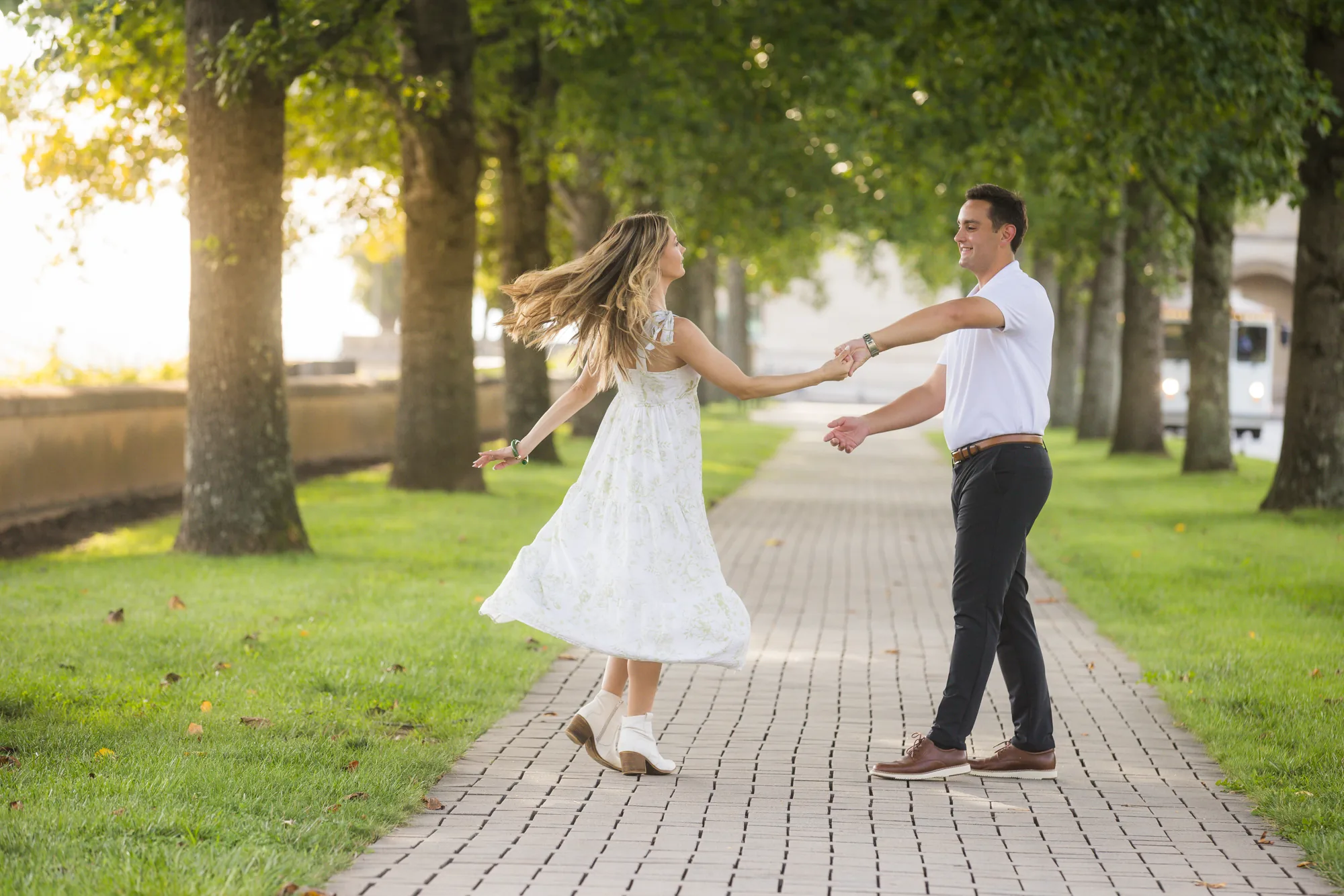 Andrea spins at arm's length from Alex in the tree-lined allée at Biltmore Estate, her white floral skirt flaring and her hair mid-swing, Alex in a white polo and dark trousers holding her hand, warm evening sun filtering through the trees, Asheville NC
