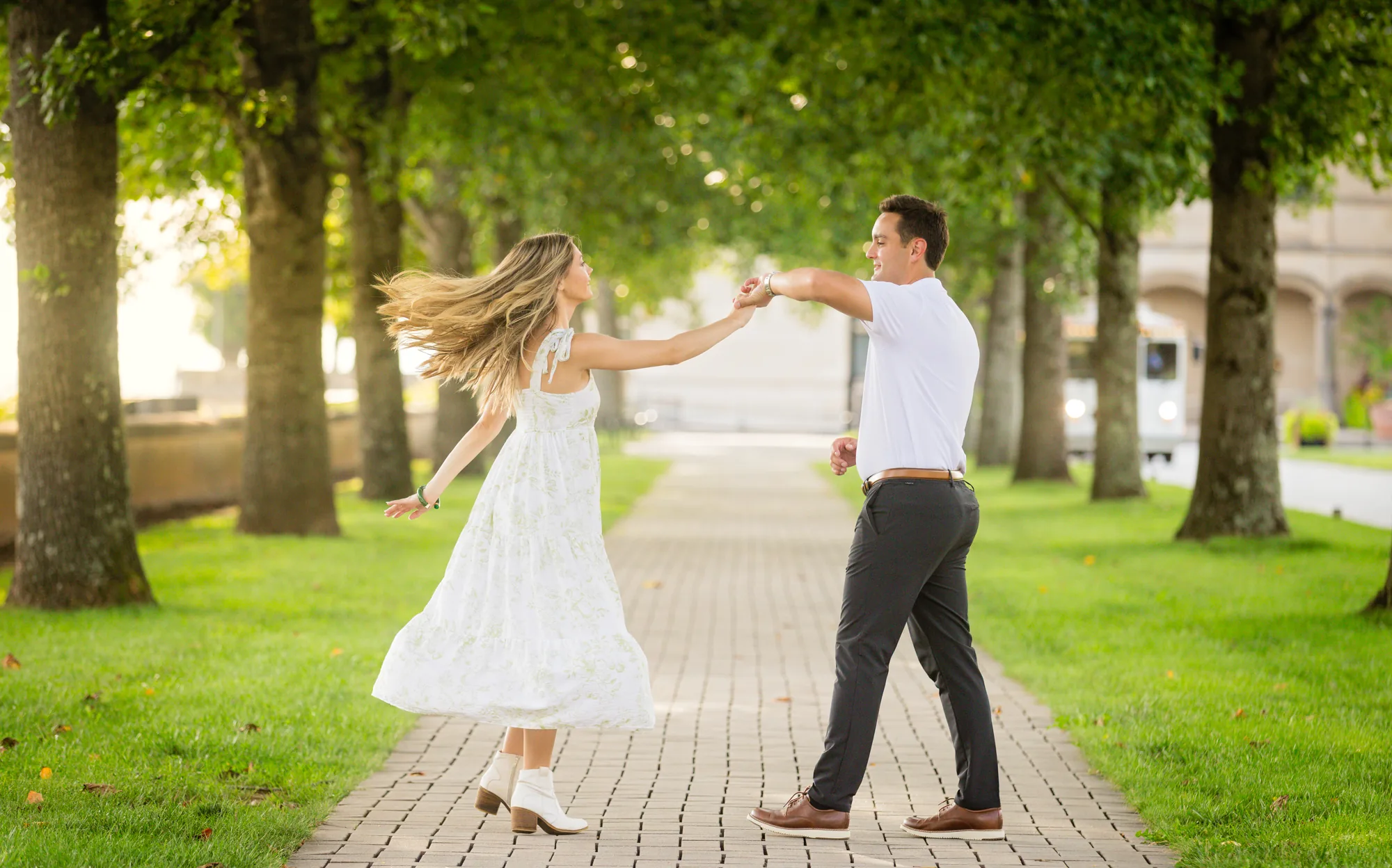Wide frame of Alex and Andrea dancing in the allée at Biltmore Estate, Andrea's dress and hair caught mid-twirl and Alex leaning back with her hand in his, the brick path running between rows of sunlit trees toward the stone of Biltmore House, Asheville NC