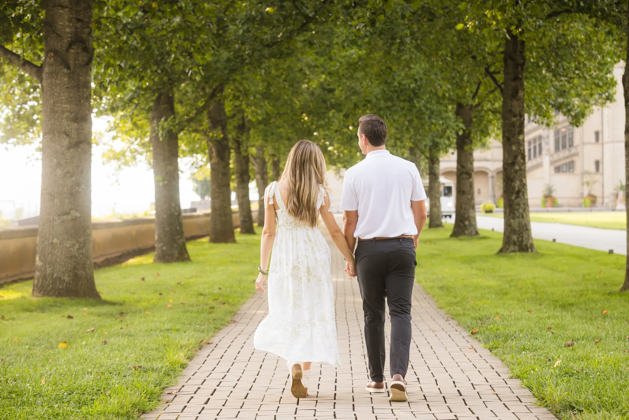 Alex and Andrea walking hand in hand away from the camera down the brick path of the tree-lined allée beside the Esplanade at Biltmore Estate, sunlit rows of trees on both sides and the stone wall of Biltmore House visible through the trunks, Asheville NC