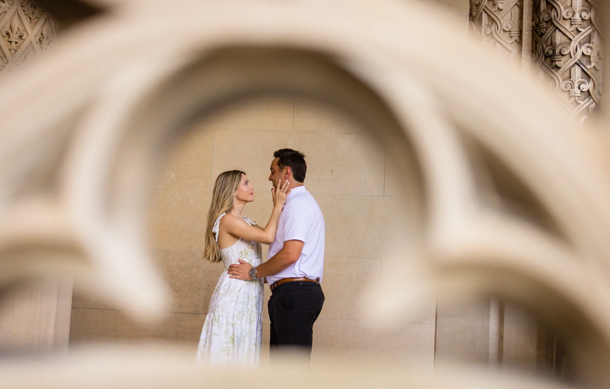 Alex and Andrea framed through the carved limestone Gothic tracery of Biltmore House at Biltmore Estate, Andrea's hand on Alex's cheek as they stand face to face against the pale stone wall, a creative fairytale engagement photo in Asheville NC