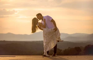 Golden hour engagement photo at Biltmore Estate: Alex dips Andrea for a kiss on the stone terrace as the sun sets in peach and gold over layered Blue Ridge Mountain ridgelines, Andrea's white floral dress and long blonde hair swinging, Asheville NC