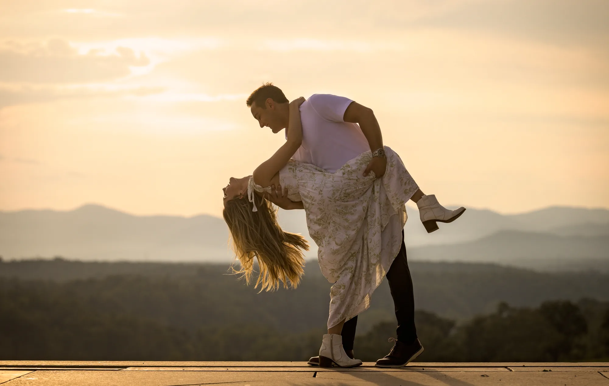 Dramatic sunset dip at Biltmore Estate in Asheville NC, Alex bending Andrea all the way back on the terrace with her hair nearly touching the stone and one white boot in the air, the Blue Ridge Mountains fading into golden haze under a glowing evening sky