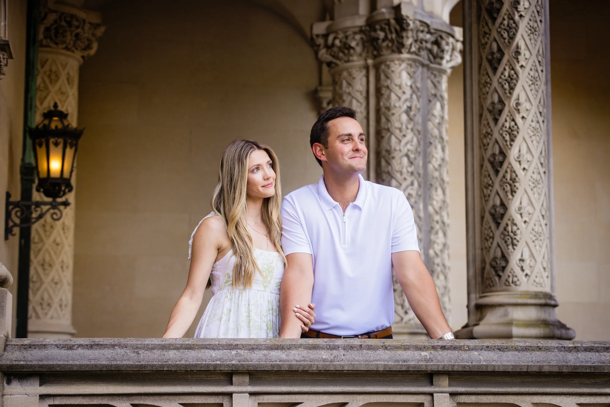 Alex and Andrea leaning on the carved stone balustrade of the front loggia of Biltmore House at Biltmore Estate, Andrea looking up at Alex beside ornate limestone columns and a glowing black iron lantern, Alex in a white polo looking off into the distance, Asheville NC