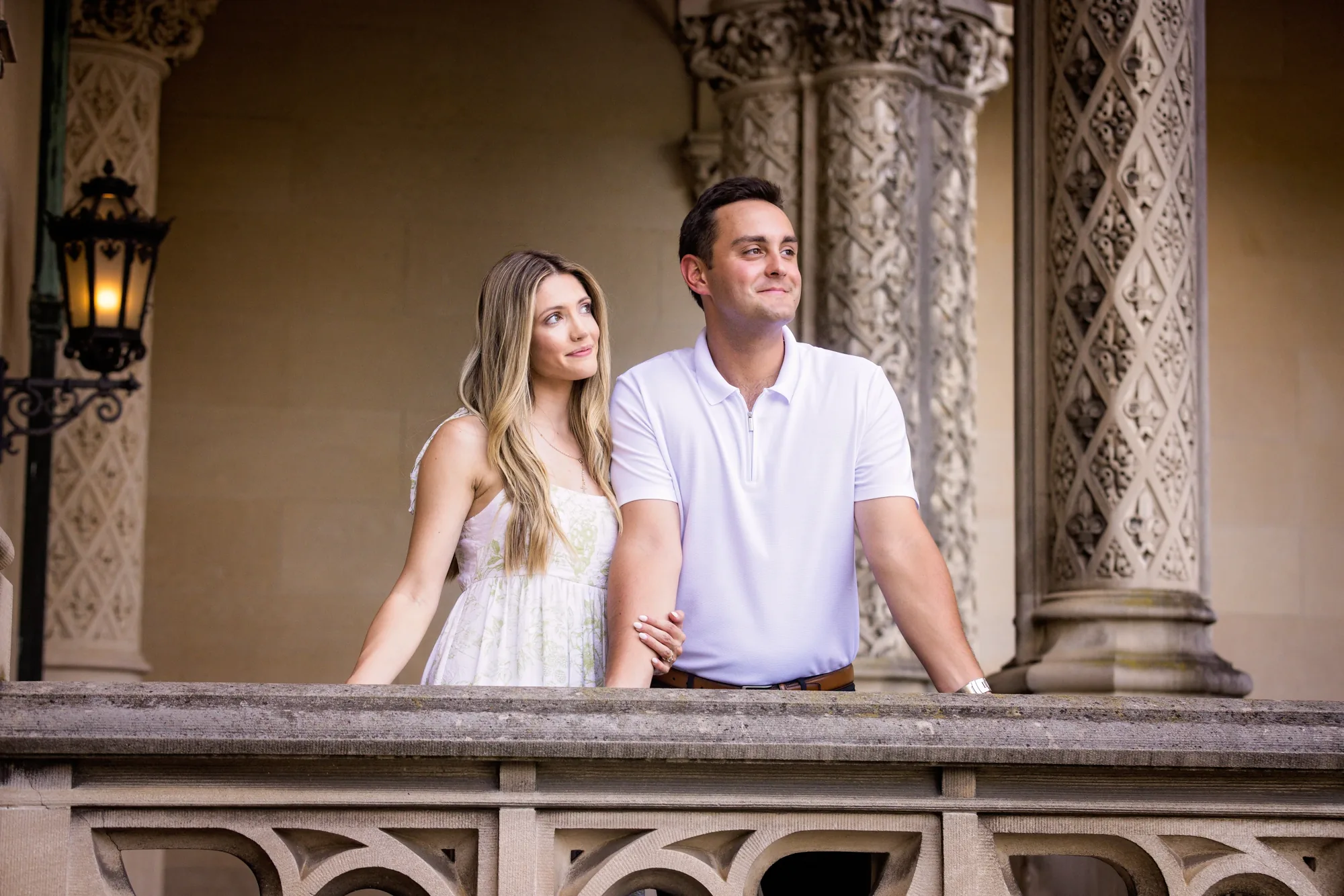 Engagement portrait of Alex and Andrea on the front loggia of Biltmore House at Biltmore Estate, Alex smiling at the camera and Andrea gazing at him, both framed by carved limestone columns, Gothic arches, and a lit iron wall lantern, Asheville NC