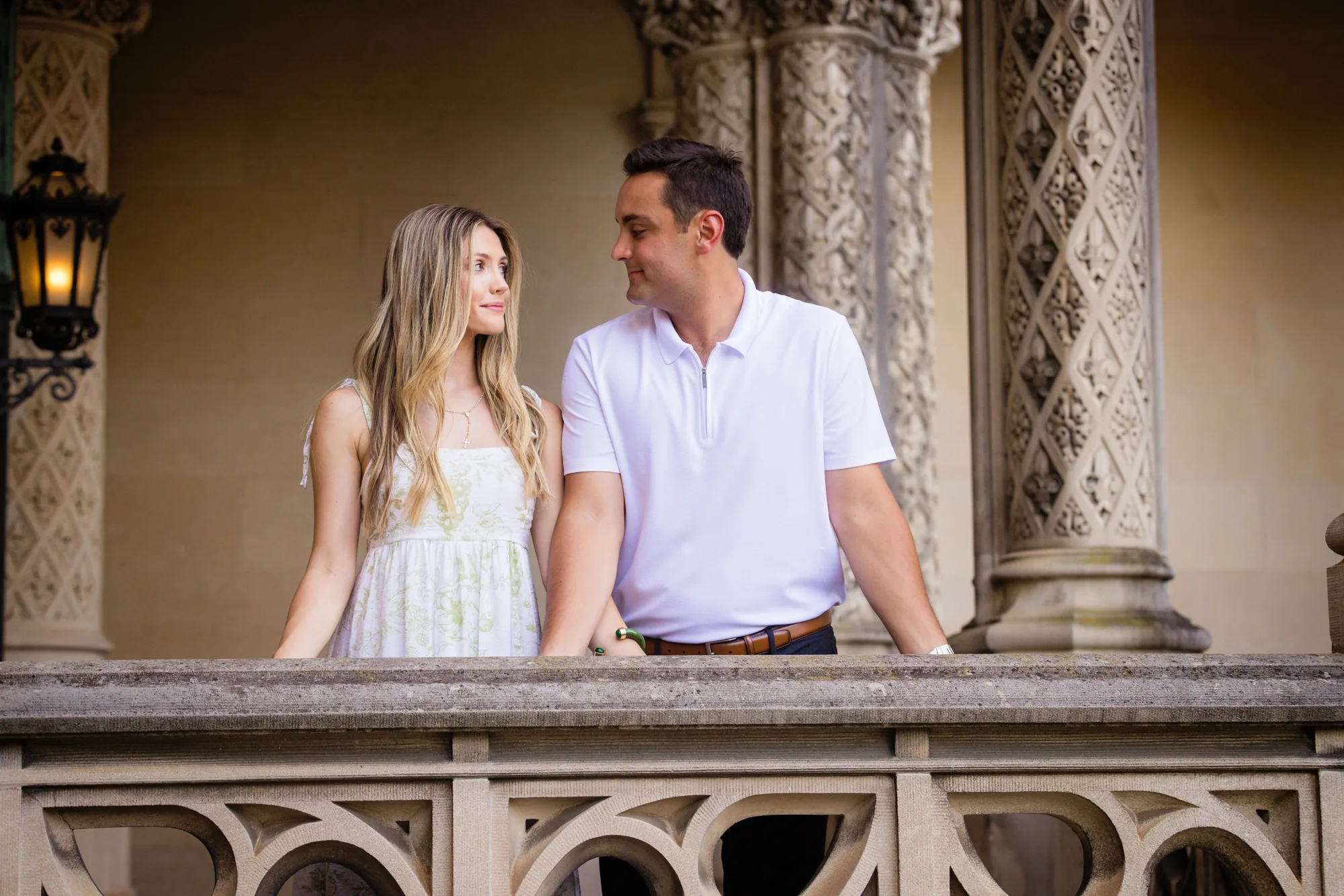 Alex and Andrea smiling at each other on the stone balustrade of the loggia at Biltmore House, Biltmore Estate, Andrea in a white floral sundress and Alex in a white zip polo with carved limestone columns and an iron lantern behind them, Asheville NC