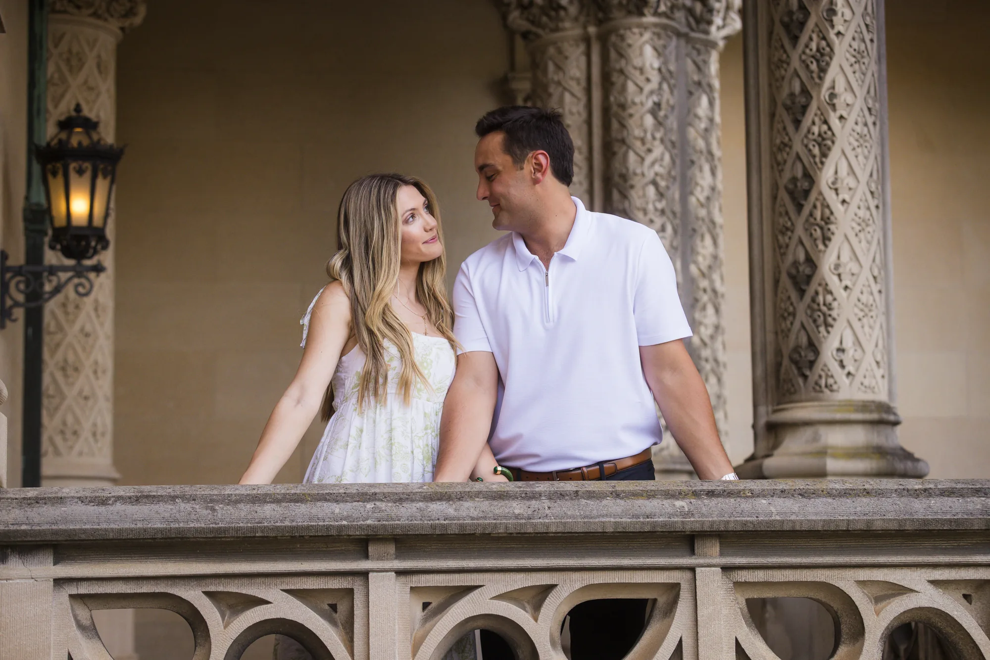 Wider frame of Alex and Andrea at the balustrade of the front loggia of Biltmore House at Biltmore Estate, the couple turned toward each other between intricately carved limestone pillars with a black iron lantern glowing to the left, Asheville NC