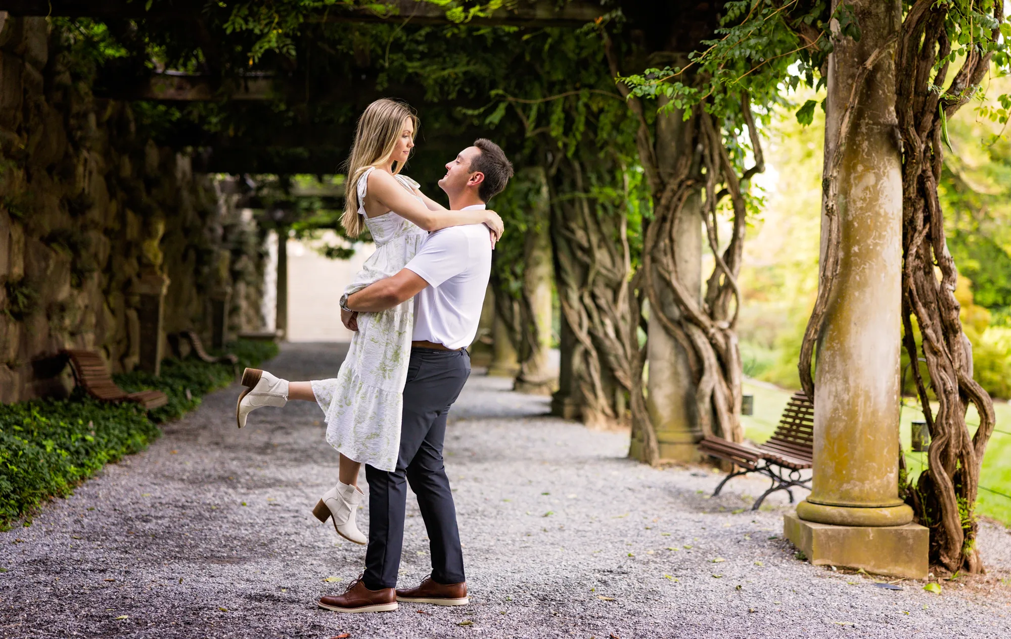 Alex lifts Andrea in the air on the gravel path of the Pergola at Biltmore Estate, Andrea laughing with one boot kicked up, surrounded by the stone columns and century-old twisting wisteria trunks of the colonnade below the Library Terrace, Asheville NC