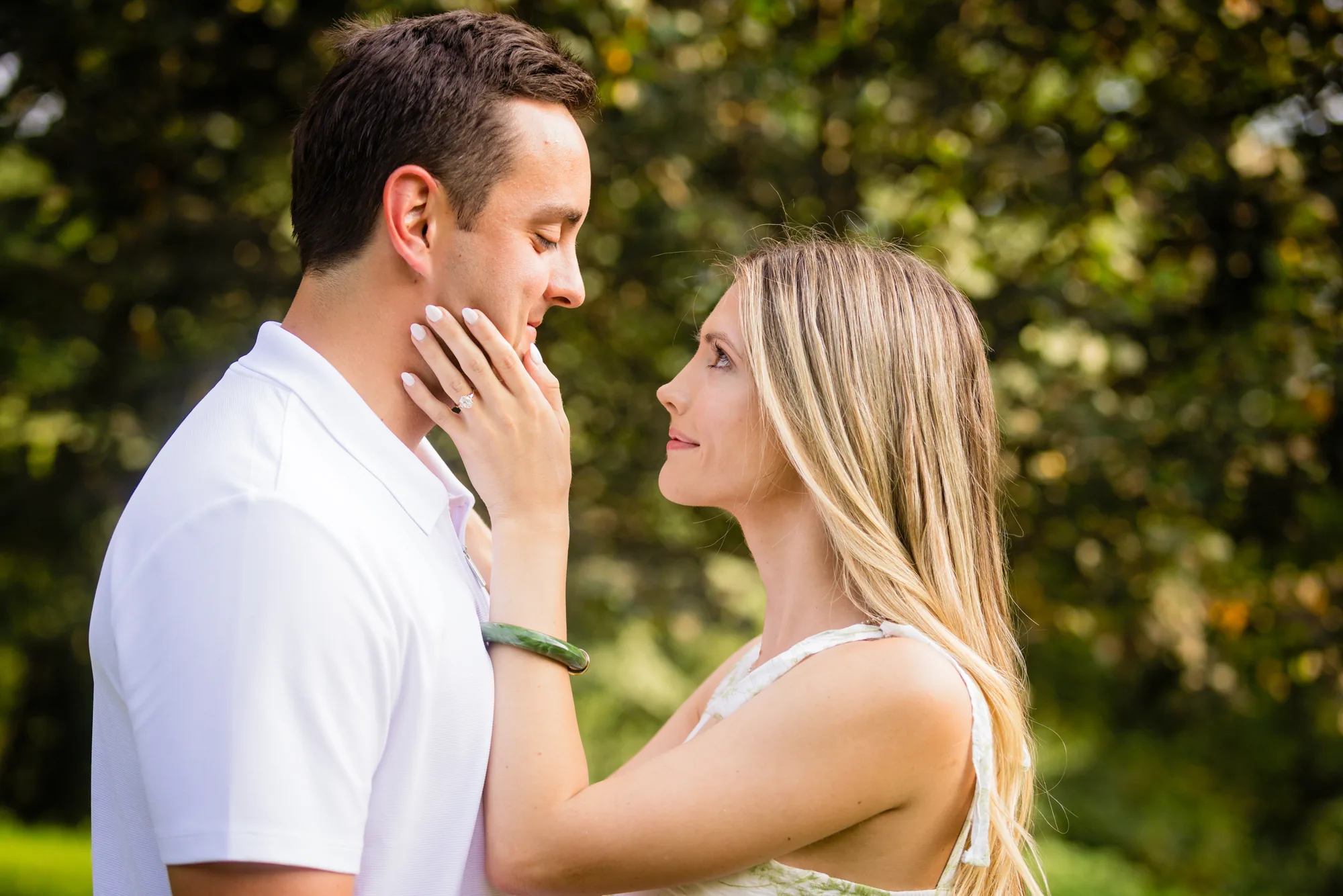 Intimate engagement portrait at Biltmore Estate in Asheville NC: Andrea cups Alex's cheek with her hand as they look into each other's eyes under a canopy of green leaves, her engagement ring and green bracelet visible, Alex in a white polo, soft dappled evening light