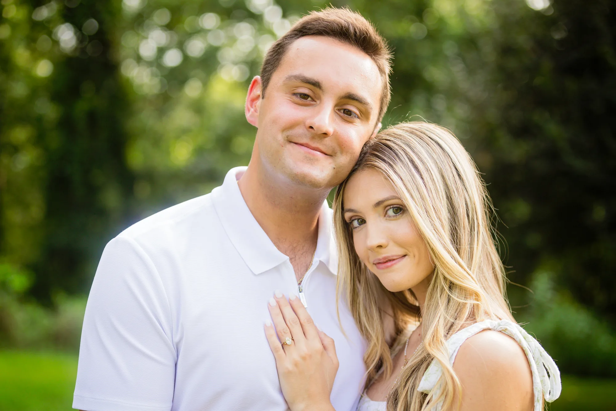 Just-engaged couple portrait at Biltmore Estate in Asheville NC: Andrea rests her head against Alex's shoulder with her new diamond engagement ring on his chest, Alex in a white zip polo and Andrea in a white floral sundress, soft sunlit green bokeh from the trees beside Diana Hill behind them