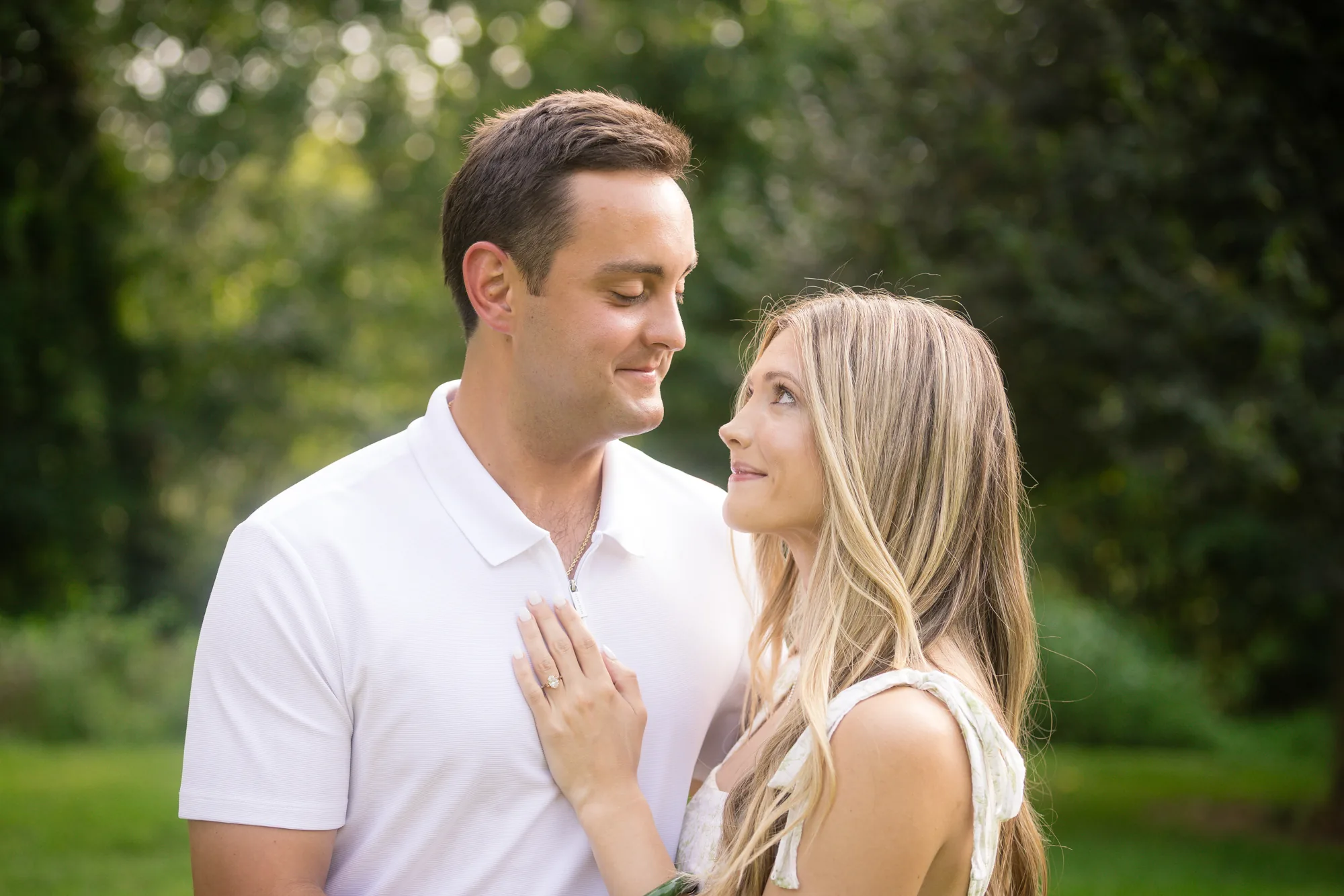 Alex and Andrea gazing at each other under the shade trees beside Diana Hill at Biltmore Estate, a romantic just-engaged portrait with Andrea's hand and engagement ring resting on Alex's white polo and dreamy backlit foliage behind them, Asheville NC