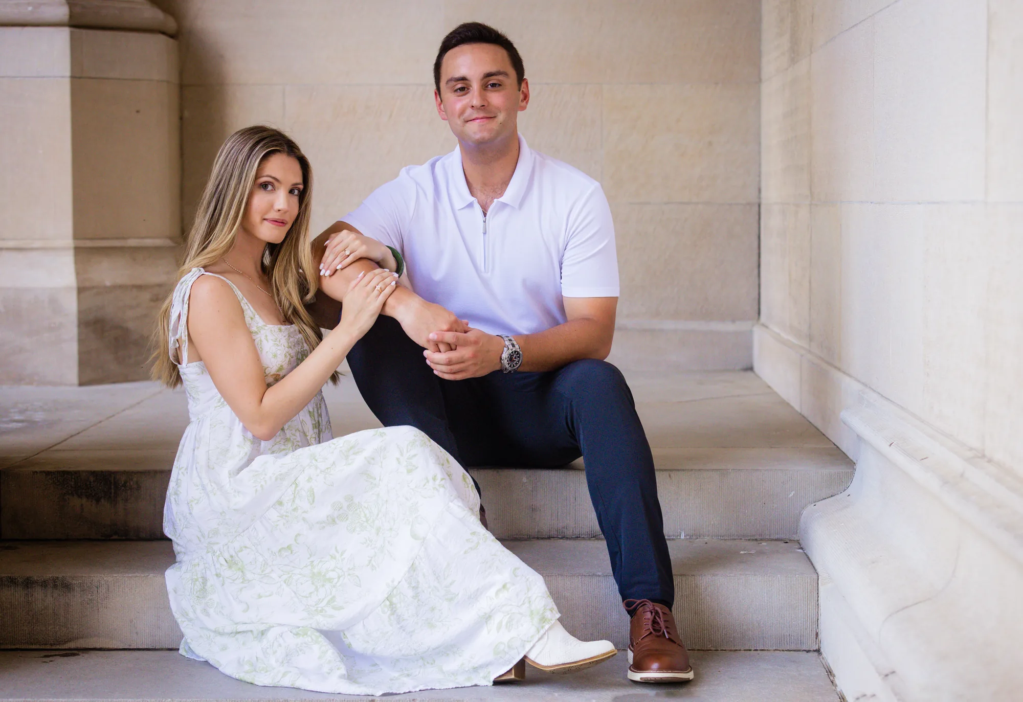 Alex and Andrea sitting together on the limestone steps of Biltmore House at Biltmore Estate, Andrea in a white and green floral dress with her hands and engagement ring resting on Alex's knee, Alex in a white polo, dark trousers, and brown leather shoes, Asheville NC