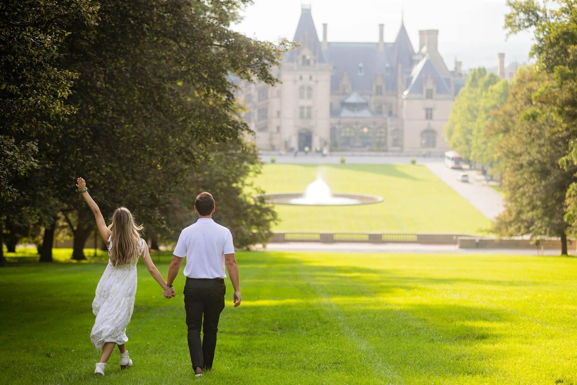 Newly engaged Andrea throws her arm in the air while walking hand in hand with Alex down the sun-drenched grass slope of Diana Hill at Biltmore Estate, the Esplanade fountain and Biltmore House glowing in golden evening light ahead of them, Asheville NC