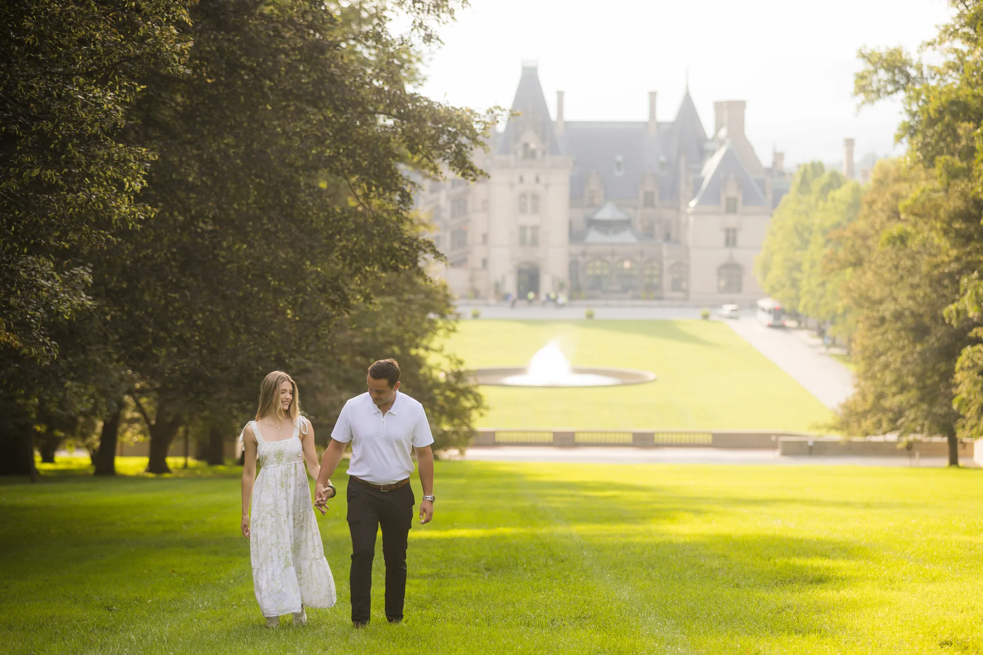 Alex and Andrea walking hand in hand toward the camera across the sunlit lawn of Diana Hill at Biltmore Estate, Andrea in a flowing white floral dress and Alex in a white polo, the towers of Biltmore House and the Esplanade fountain hazy and backlit behind them