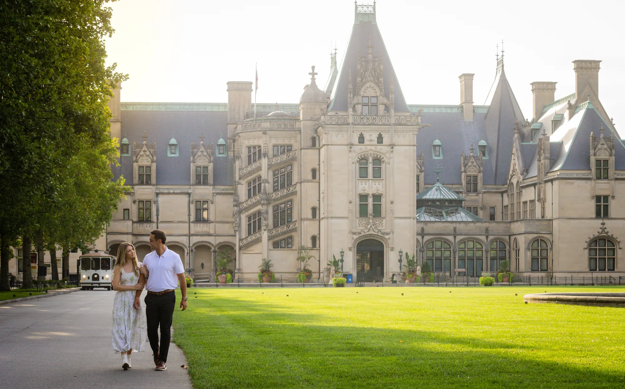 Alex and Andrea walking hand in hand along the edge of the front lawn of Biltmore House at Biltmore Estate, the full limestone facade with its steep slate roofs, spires, and turrets rising behind them under a bright late-summer sky in Asheville NC