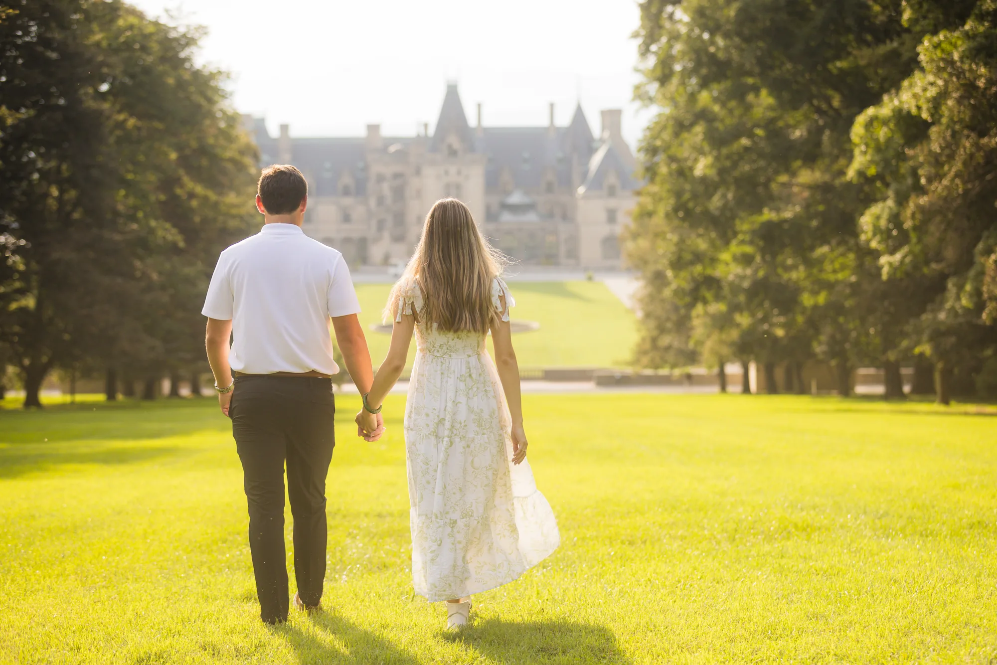 Alex and Andrea photographed from behind walking hand in hand down Diana Hill toward Biltmore House at Biltmore Estate, the couple in white with the Esplanade fountain and the limestone mansion framed by tall trees in warm sunlit haze, Asheville NC
