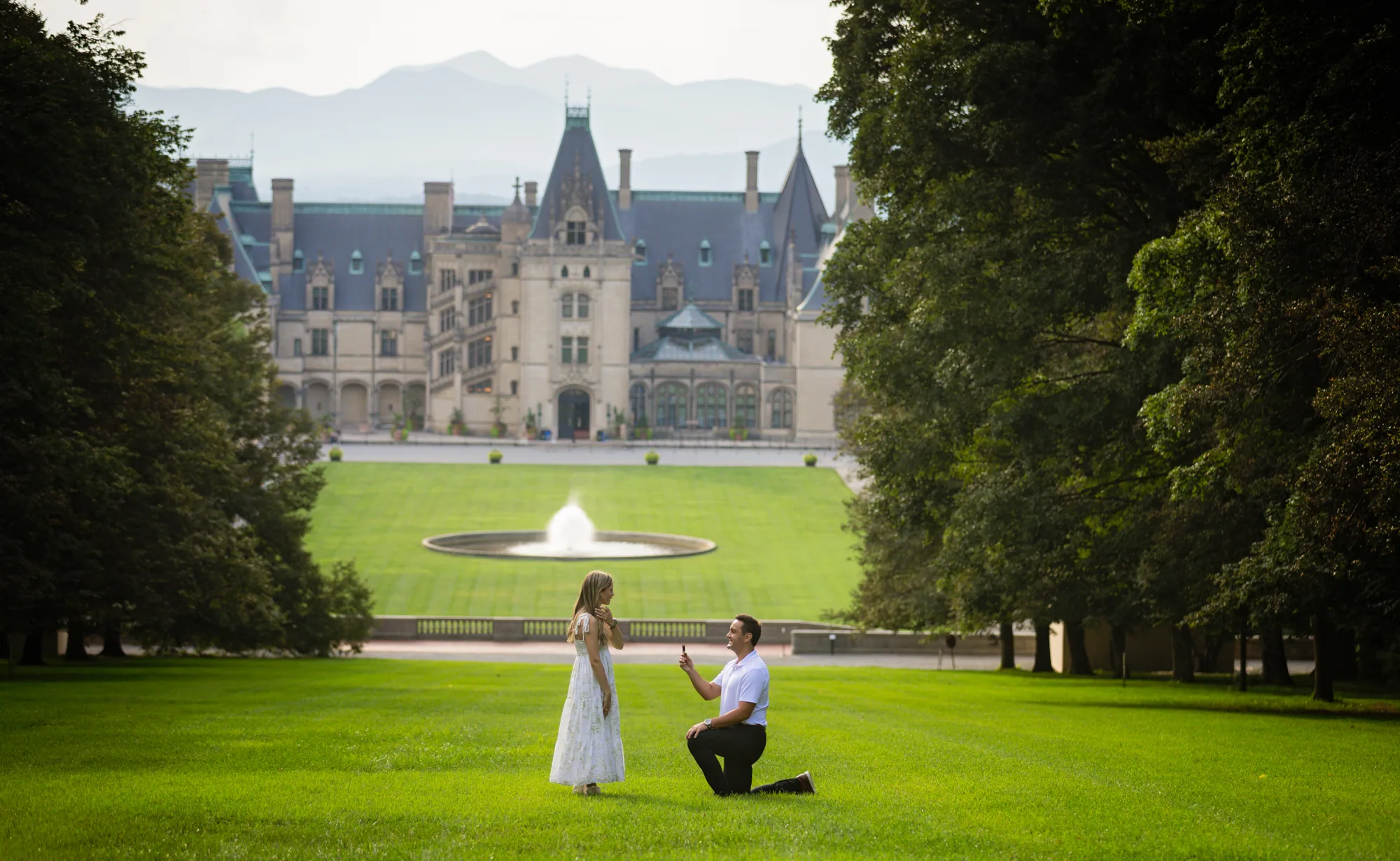 Surprise Biltmore Estate proposal on Diana Hill in Asheville NC: Alex kneels on one knee in a white polo and dark trousers holding up an engagement ring to Andrea, who stands in a white floral sundress on the sunlit grass slope above the Esplanade, with the fountain pool, the limestone facade of Biltmore House, and the Blue Ridge Mountains behind them