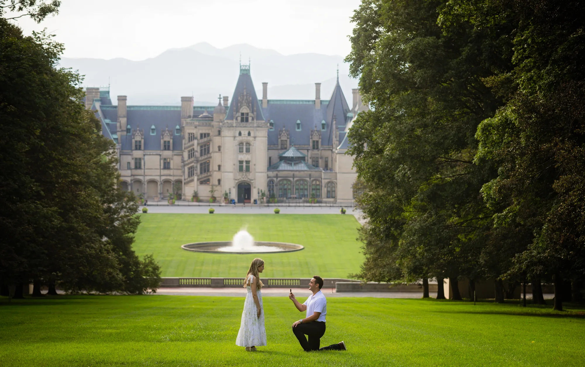 Wide view of a surprise marriage proposal at Biltmore Estate, Alex kneeling before Andrea on the green lawn of Diana Hill framed by tall trees, the Esplanade fountain and the full width of Biltmore House and hazy Blue Ridge ridgelines in the background on a summer evening in Asheville NC