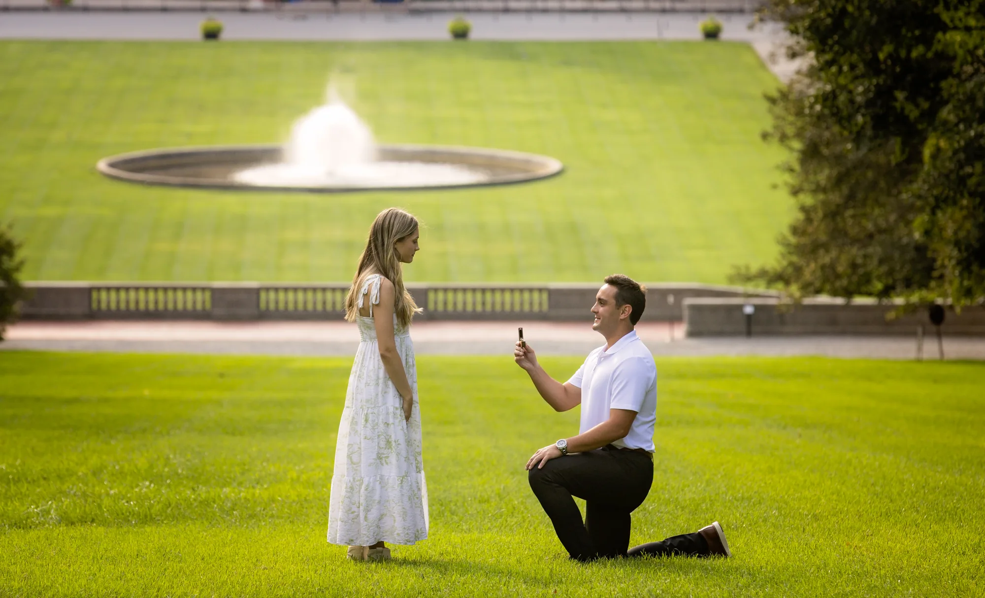 Close telephoto view of Alex proposing to Andrea on Diana Hill at Biltmore Estate, Alex on one knee in a white polo holding the open ring box while Andrea in a white and green floral dress reacts, the Esplanade fountain spraying and the stone balustrade of the Rampe Douce behind them in warm evening light