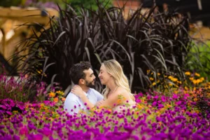 Ryan and Amanda sitting close together among vivid magenta flowers and orange blooms in the Walled Garden at Biltmore Estate, gazing at each other, dark purple ornamental grasses rising behind them