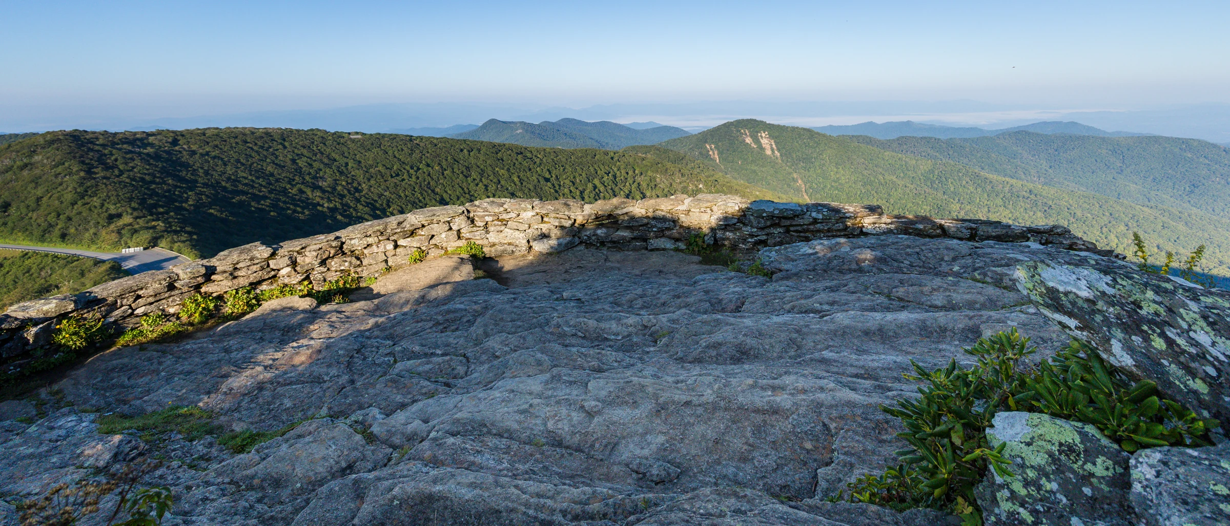 A broad rock viewing platform bordered by a low stone wall at the lower overlook on Craggy Pinnacle, with mountain ridges beyond