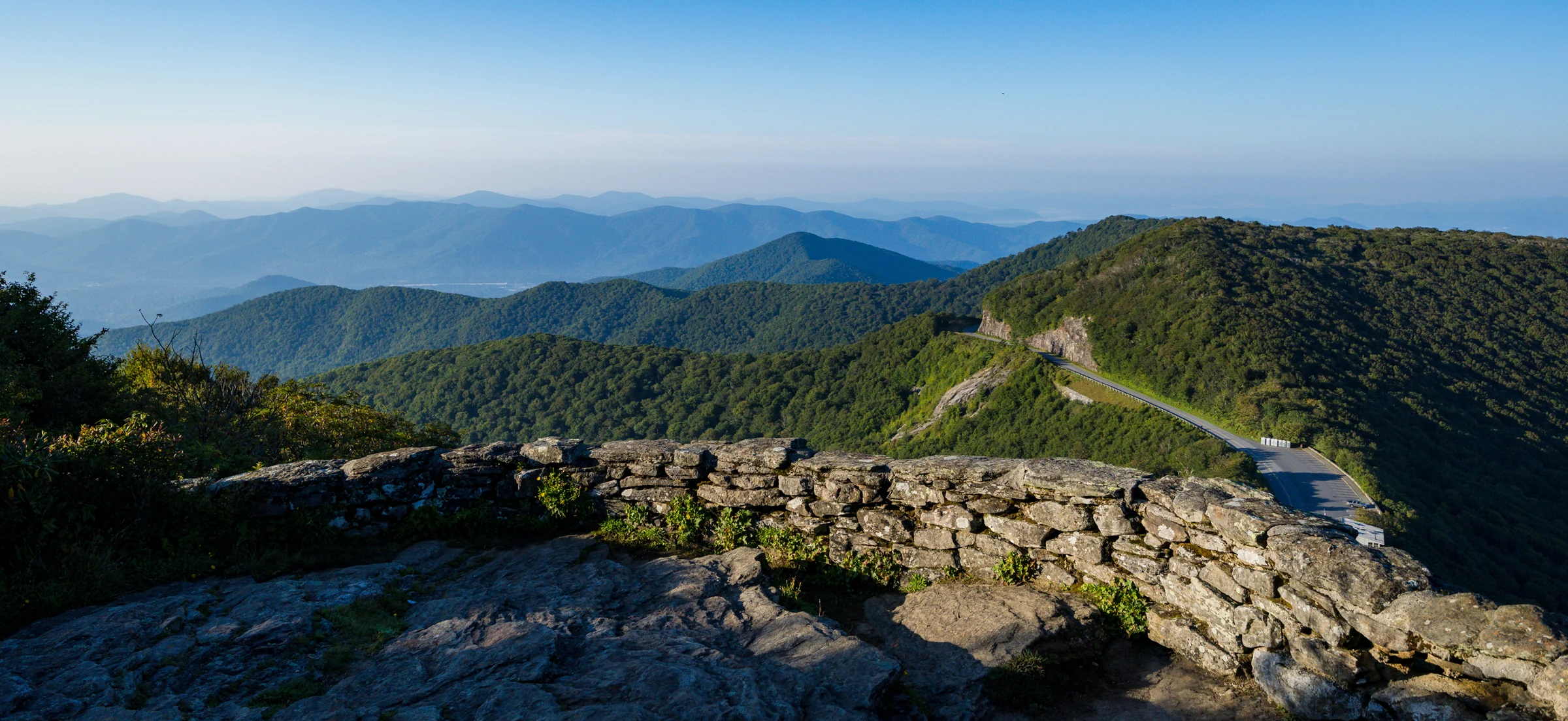 The Blue Ridge Parkway curves along a green mountainside below the stone overlook on Craggy Pinnacle, with the Great Craggy Mountains beyond