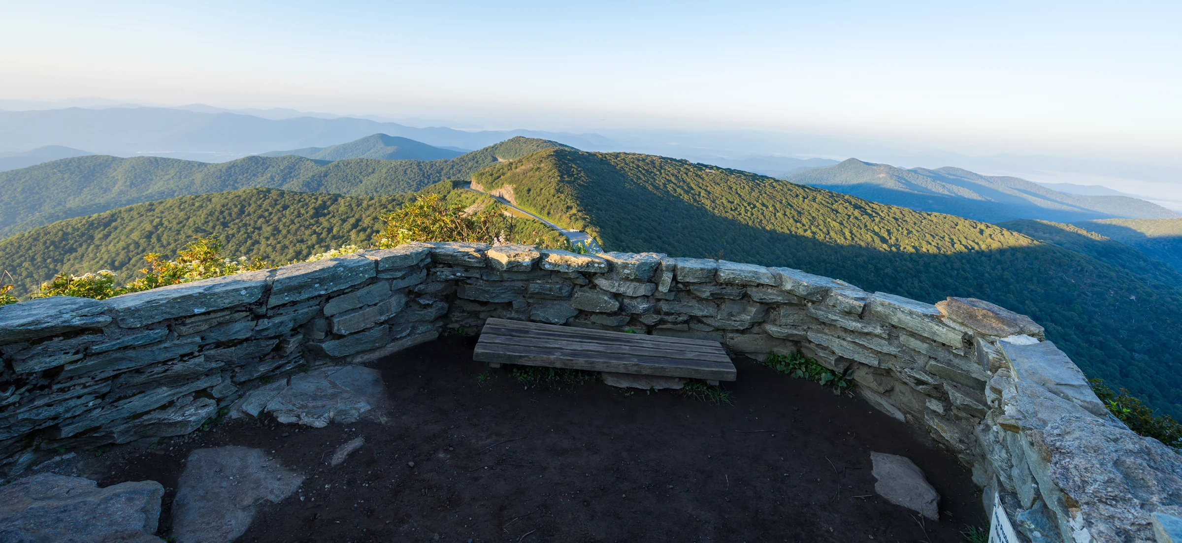 The stone-walled summit overlook and bench on Craggy Pinnacle in clear morning light, with ridges of the Black Mountains and Great Craggy Mountains beyond