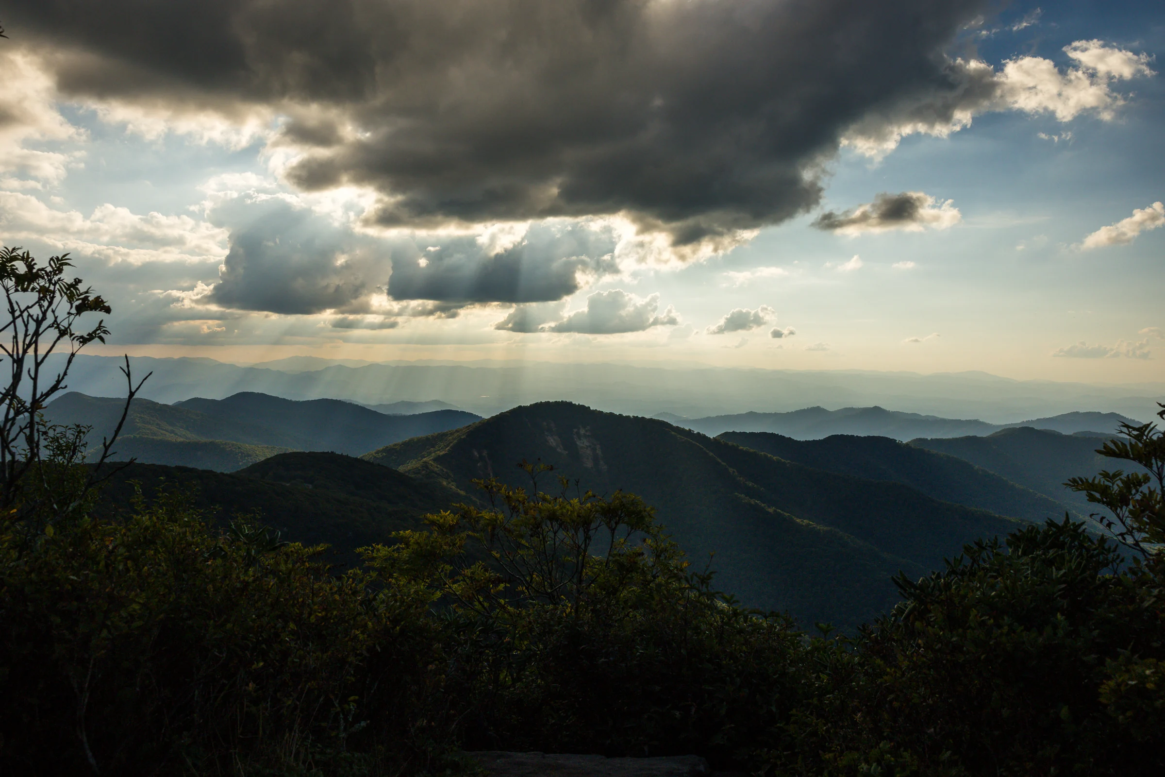 Sunbeams break through dark clouds over layers of blue mountains at sunset, seen from the west ledge on Craggy Pinnacle on the Blue Ridge Parkway