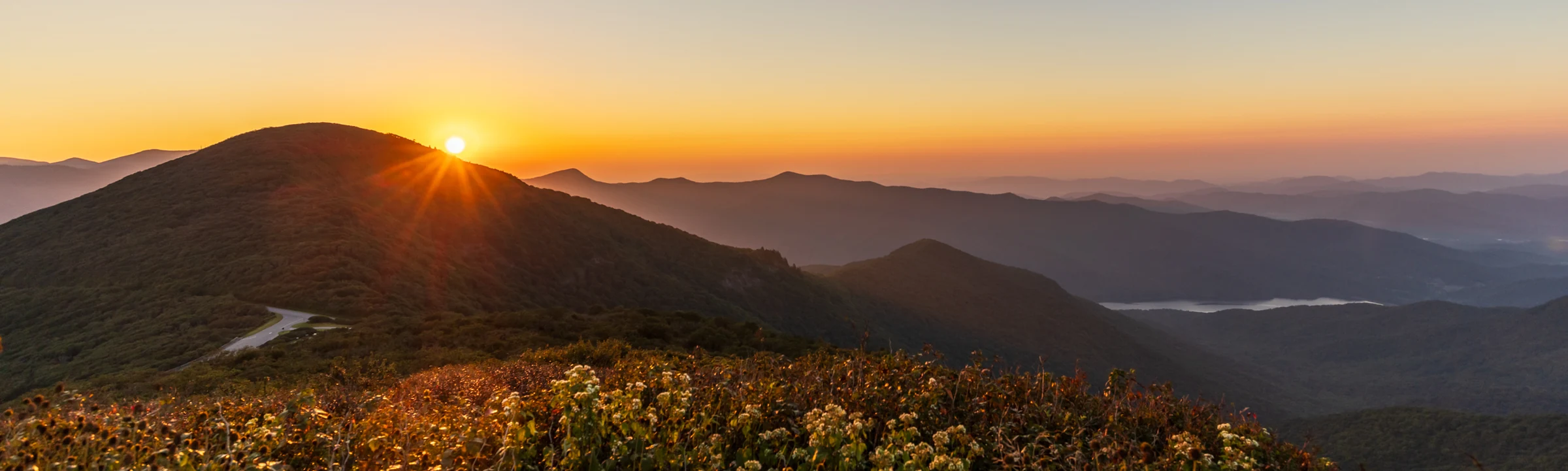 The rising sun breaks over a mountain ridge in a wide panorama from the summit of Craggy Pinnacle