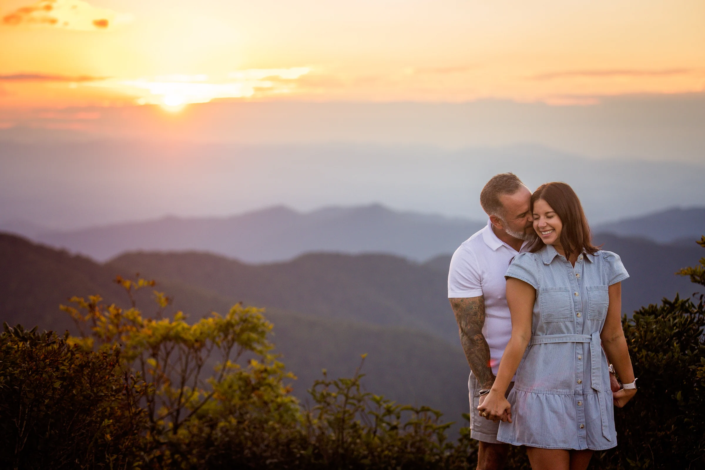 Scott kisses Jaci's cheek beneath a bright sunset sky with layered blue mountain ridges behind them at Craggy Pinnacle
