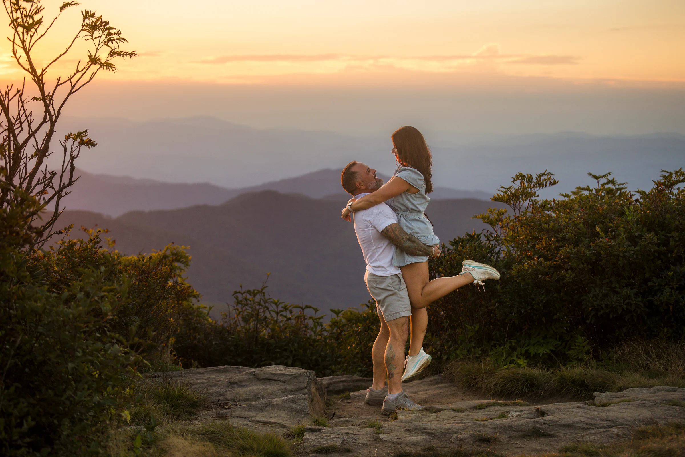 Scott lifts Jaci off the rock on the west ledge of Craggy Pinnacle with the Blue Ridge Mountains behind them at sunset