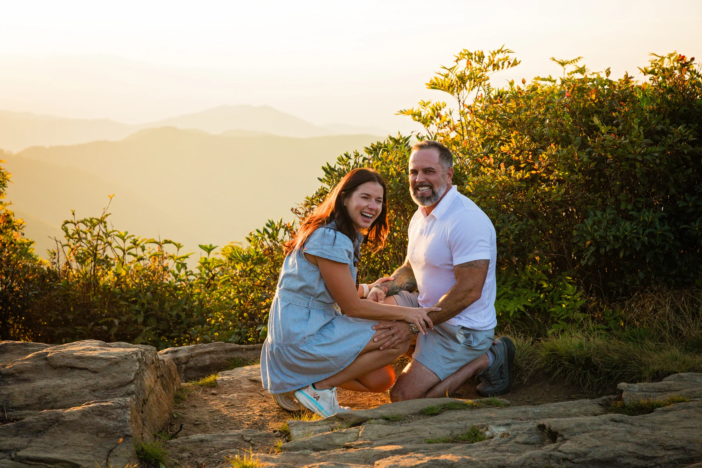 Scott and Jaci laughing together seconds after the proposal on the west ledge of Craggy Pinnacle, sunset color on the Blue Ridge Mountains behind them