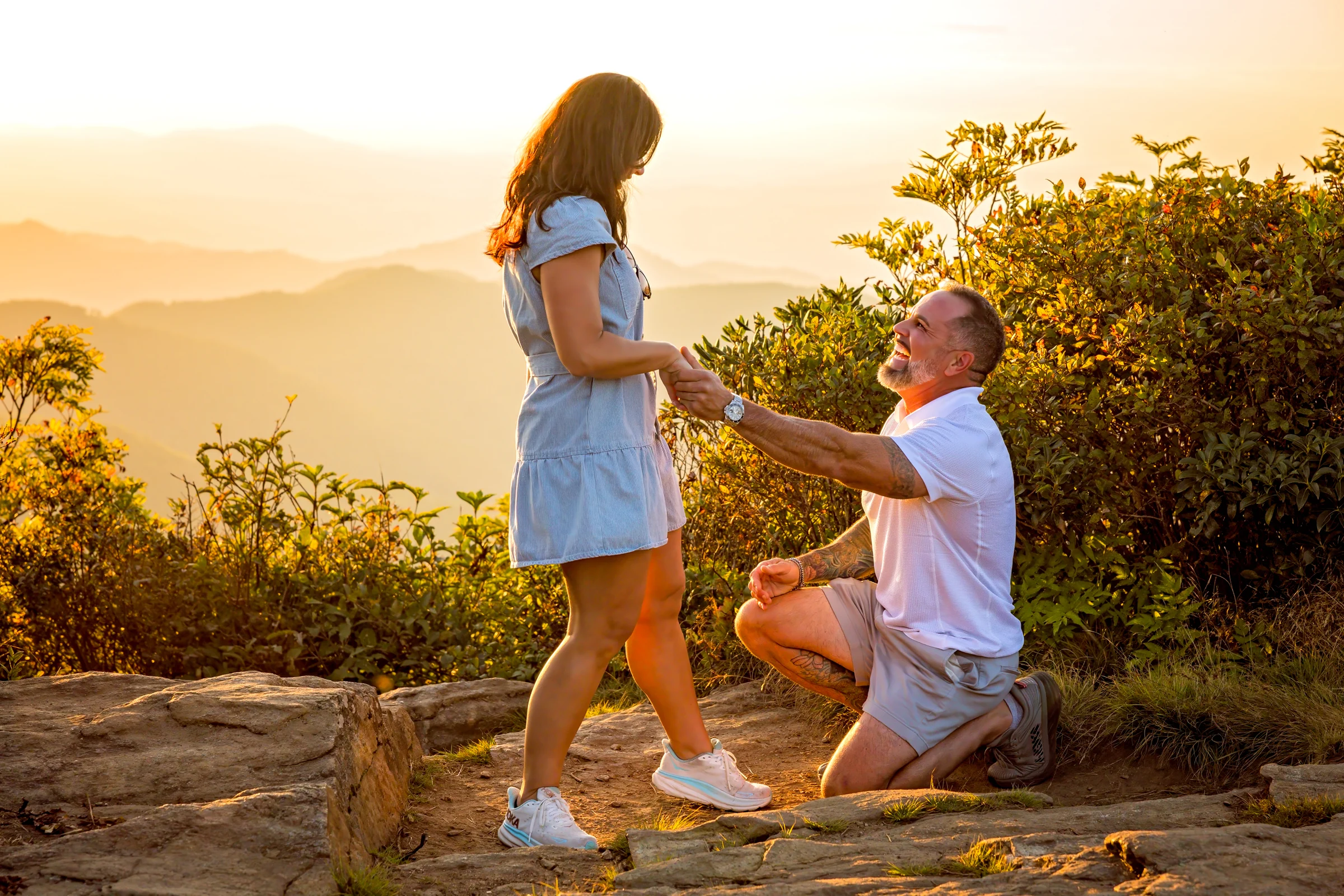 Scott holds up the engagement ring to Jaci on the west ledge of Craggy Pinnacle at sunset, mountains fading into the haze behind them