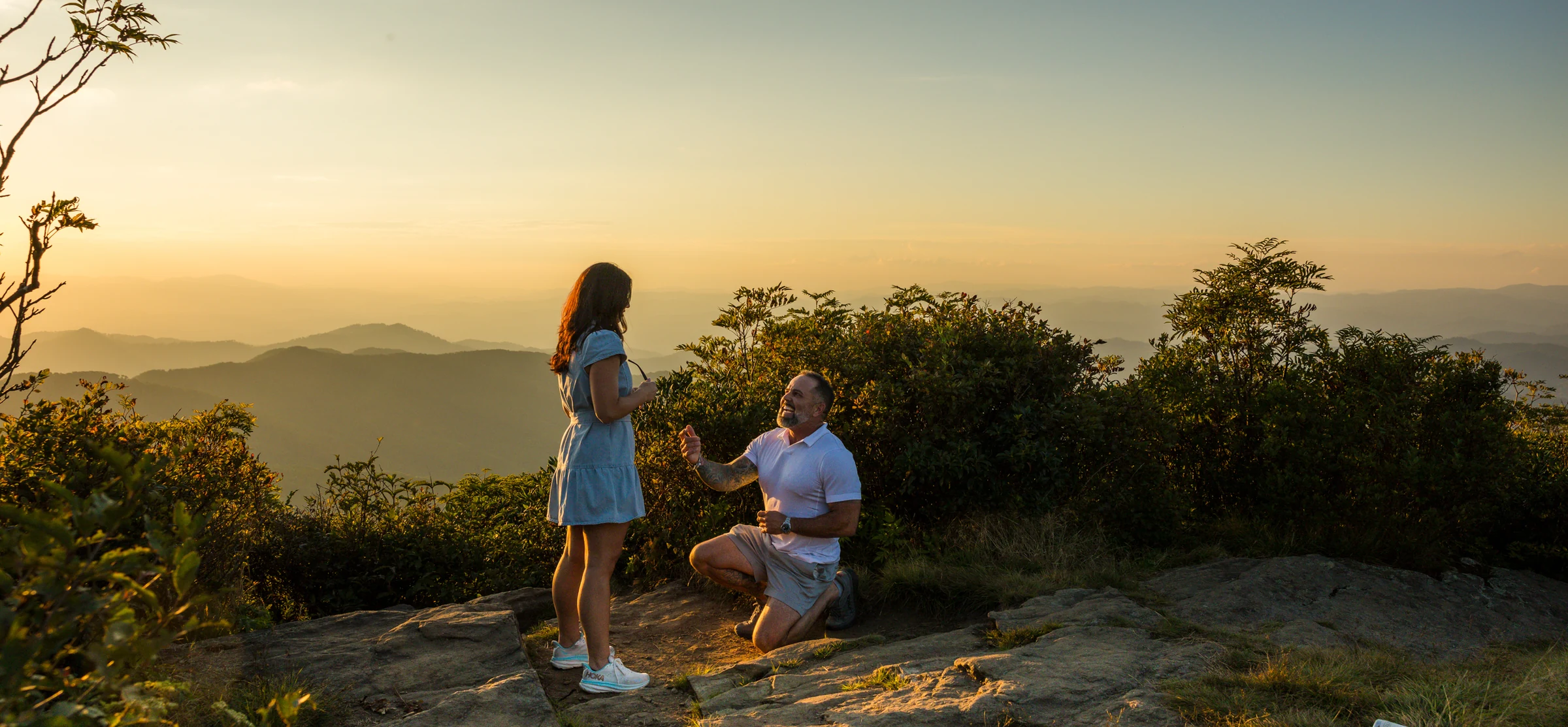 Scott kneels to propose to Jaci on the west ledge of Craggy Pinnacle with golden sunset light on the mountain ridges behind them