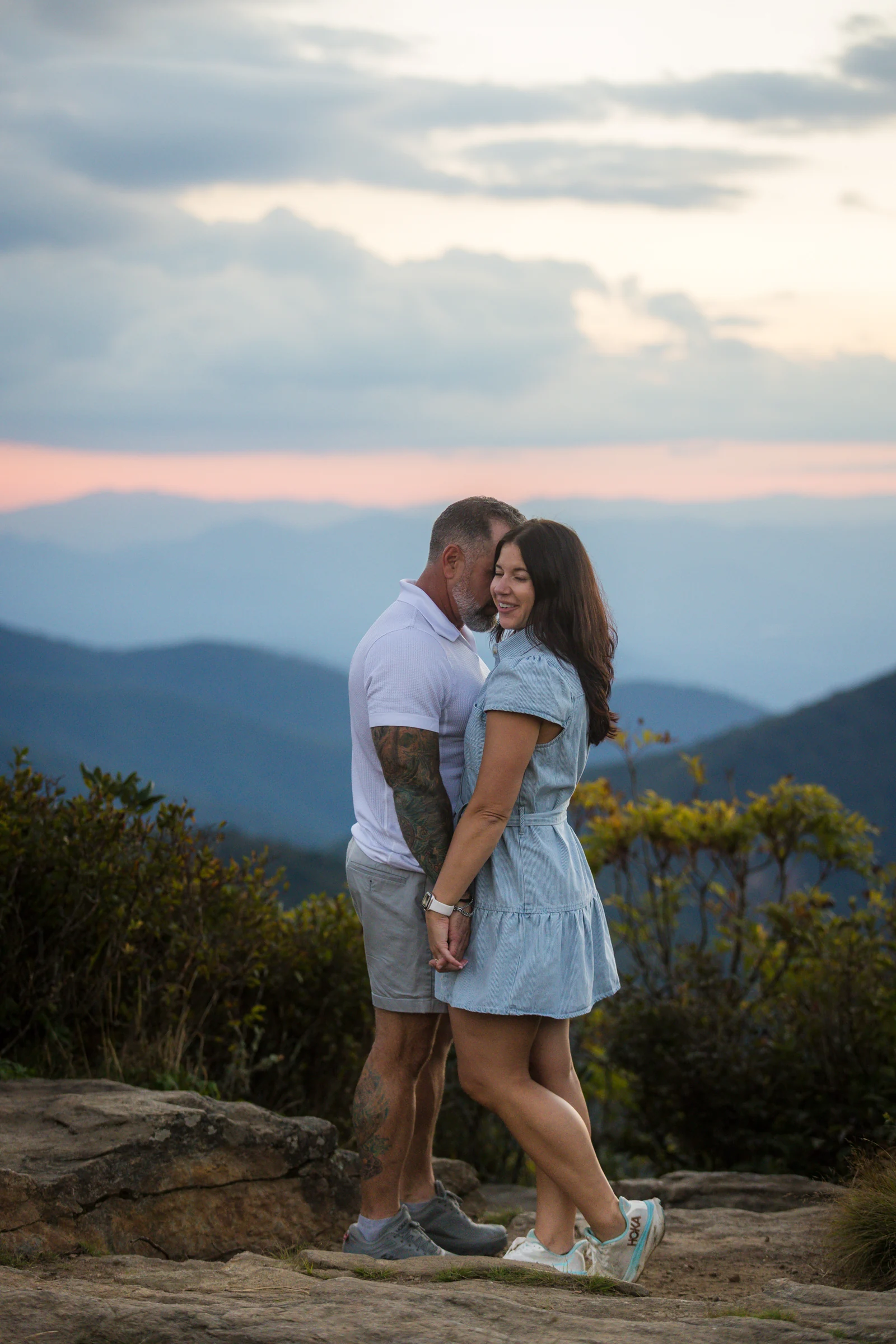 A vertical portrait of Scott and Jaci embracing on the west ledge of Craggy Pinnacle with mountains fading into the distance