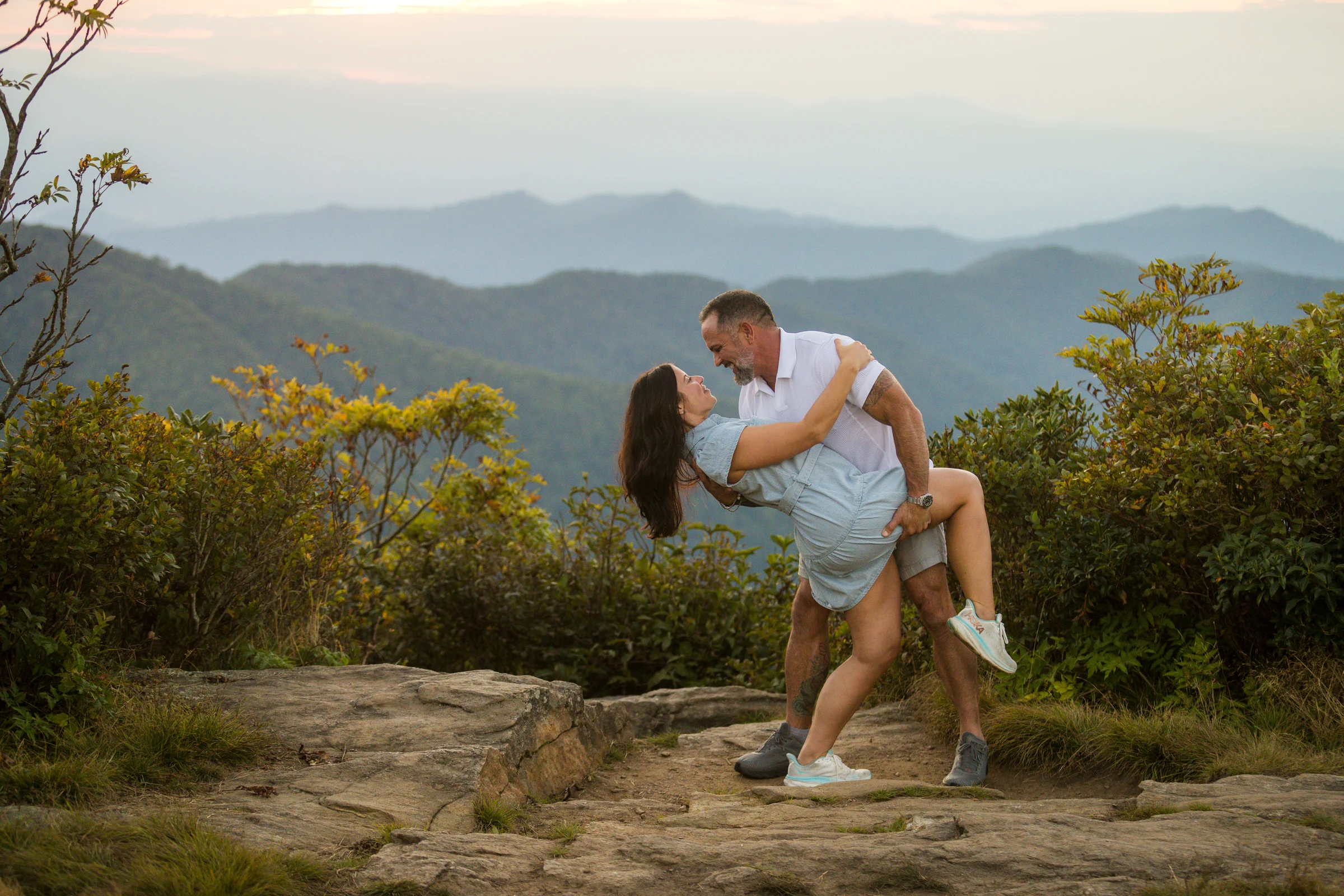 Scott lifts Jaci in a wide blue-hour portrait on Craggy Pinnacle with the ridgelines behind them